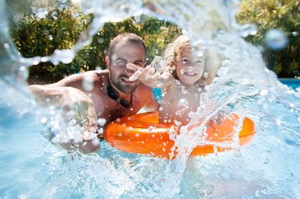 Vater und Kind spielen lachend im Pool, während Wasser spritzt, das Kind sitzt in einem orangen Schwimmring