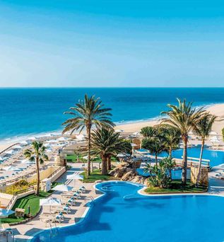 Großer Hotelpool mit Palmen und Liegestühlen, dahinter Sandstrand und blauer Ozean unter klarem Himmel