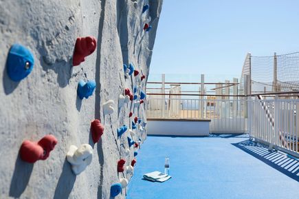 Boulder‑Wand auf der AIDAcosma mit farbigen Klettergriffen auf einem sonnigen Outdoor‑Deck, daneben eine Matte und eine Wasserflasche auf blauem Bodenbelag
