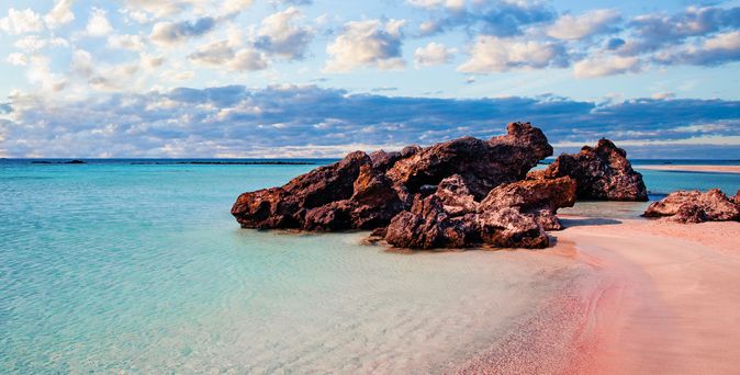 Rosa Sandstrand mit sanften Wellen und großen Felsen im Wasser, unter einem Himmel mit Wolken. Aufnahme von Kreta.