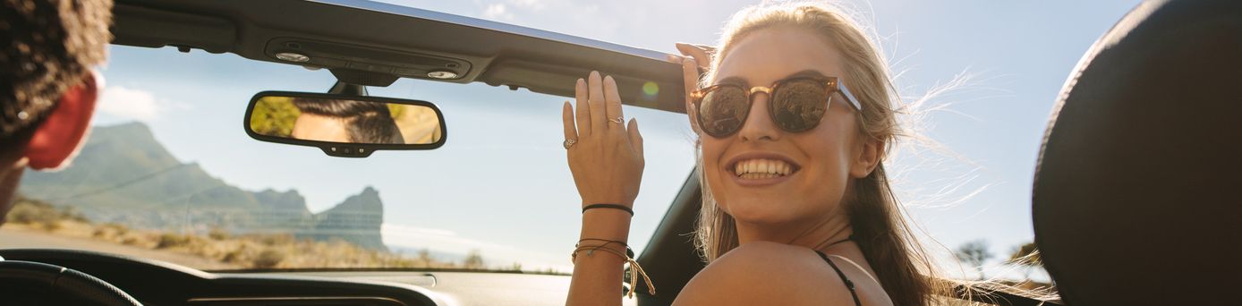 Ein offenes Auto mit zwei Personen im Innenraum. Die Person auf dem Beifahrersitz trägt ein gestreiftes Oberteil und hat die Hand erhoben. Im Hintergrund sind Berge und ein sonniger Himmel sichtbar.
