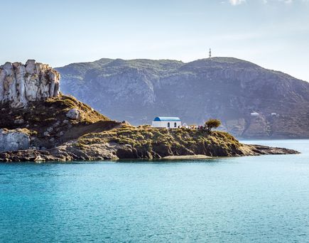 Kleine felsige Insel mit weiß-blauem Gebäude im Meer vor bewaldetem Berg im Hintergrund
