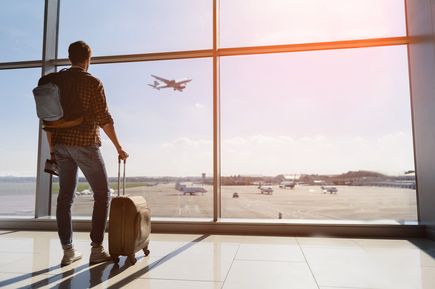 Reisender steht mit Koffer und Rucksack am Flughafen vor einer großen Glasfront und blickt auf startende und parkende Flugzeuge auf dem Rollfeld