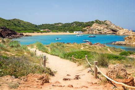 Sandweg führt zur Bucht Cala Pregonda auf Menorca mit goldgelbem Sand, rötlichen Felsen und Booten im klaren Wasser