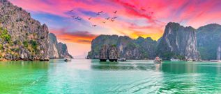 Kalksteinfelsen und Ausflugsboote in der Halong-Bucht in Vietnam auf ruhigem Wasser unter farbigem Abendhimmel