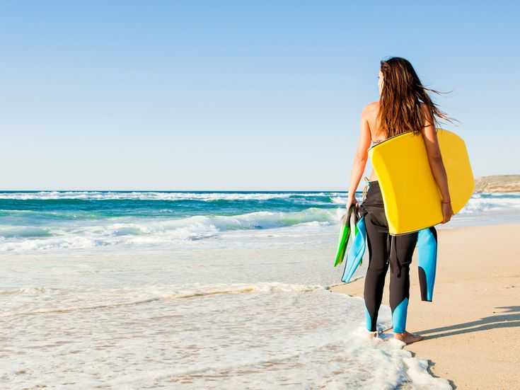 Frau im Neoprenanzug steht mit Bodyboard und Flossen am Strand und blickt auf das Meer mit Wellen