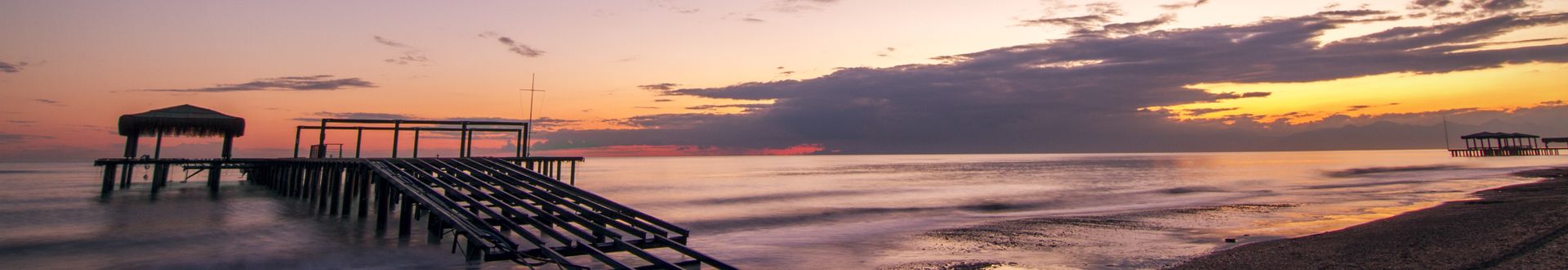 Verlassener Holzsteg mit Strohdach am Strand von Belek in der Türkei bei Sonnenuntergang, ruhiges Meer und violett-orange gefärbter Himmel