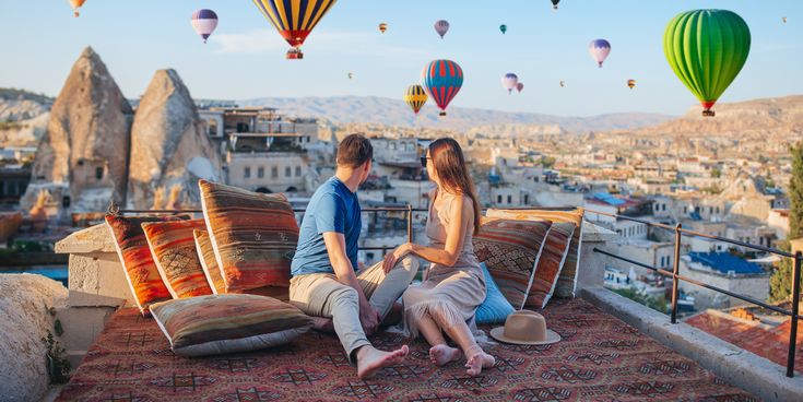 Paar sitzt auf einer Terrasse in Kappadokien, Türkei, mit Blick auf Felsen und zahlreiche Heißluftballons am Himmel