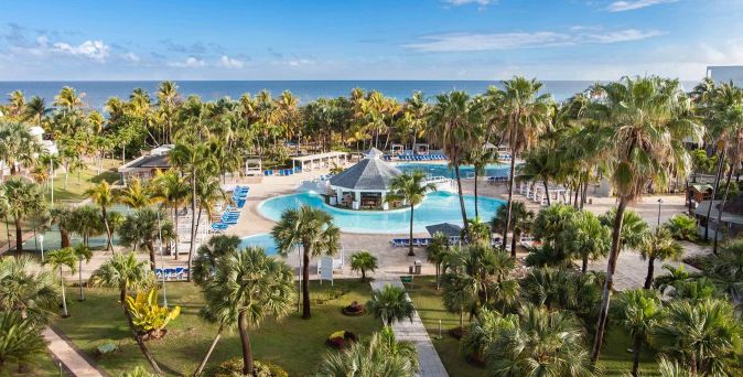 Hotelpool mit Bar in der Mitte, umgeben von Palmen und Liegestühlen, im Hintergrund das Meer und blauer Himmel