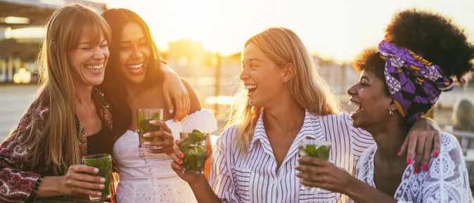 Vier Freundinnen lachen und stoßen bei Sonnenuntergang mit Cocktails an, während sie Zeit im Freien genießen