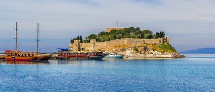 „Historische Festung auf der Taubeninsel vor Kusadasi mit klar blauem Meer, traditionellen Ausflugsbooten und grüner Küstenlandschaft bei sonnigem Wetter