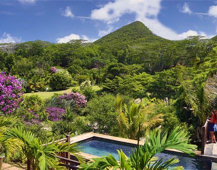 Blick auf tropische Gartenanlage mit blühenden Büschen, Palmen, Pool und bewaldetem Berg im Hintergrund, zwei Personen auf Terrasse