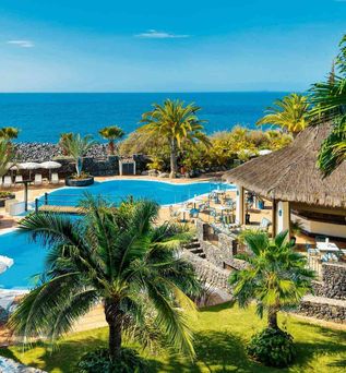 Hotelpool mit Liegestühlen und Sonnenschirmen, umgeben von Palmen und einer Strohdachbar, im Hintergrund das Meer unter blauem Himmel.