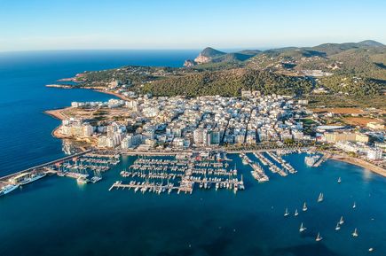 Luftaufnahme von San Antonio auf Ibiza mit dicht bebauter Küstenstadt, Yachthafen voller Boote und umliegenden grünen Hügeln