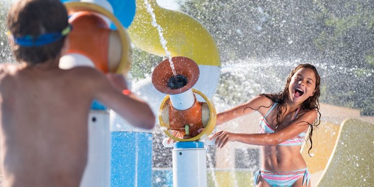 Zwei Kinder spielen lachend im Splash-Pad, Wasserfontänen und Spritzgeräte sorgen für Abkühlung in einer Ferienanlage
