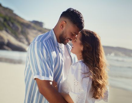 Junges Paar steht umarmt am Strand, lächelt sich an, im Hintergrund Meer und Küste bei sonnigem Wetter