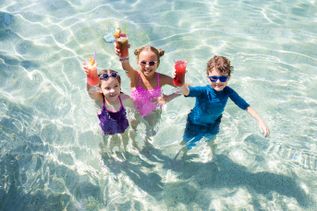 Vier Kinder stehen im flachen Pool und halten farbenfrohe Getränke fröhlich in die Höhe, während Sonnenlicht funkelnde Reflexionen auf der Wasseroberfläche erzeugt