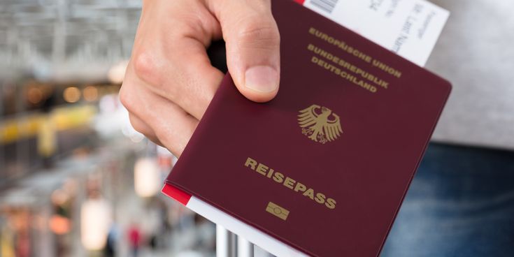 Eine Hand hält einen deutschen Reisepass in Rot mit dem Bundesadler und dem Text "Reisepass". Im Hintergrund sind unscharfe Flughafen-Elemente zu sehen.
