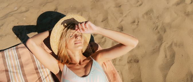 Person liegt entspannt auf einem gestreiften Handtuch im warmen Sand und hält einen Strohhut über dem Gesicht, während die Sonne die Strandfläche beleuchtet