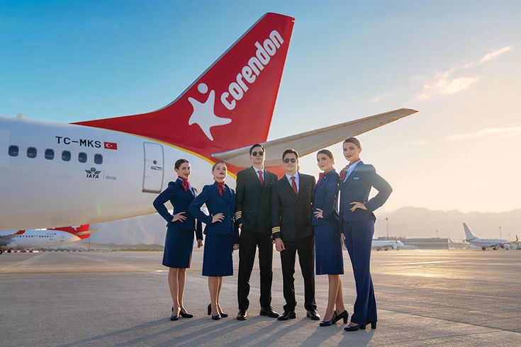 Flugpersonal der Corendon Airlines steht in Uniform auf dem Vorfeld vor dem Leitwerk einer Boeing 737, Corendon-Logo deutlich sichtbar, Flughafen und weitere Flugzeuge im Hintergrund