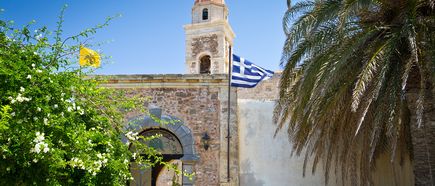 Steinerner Kirchturm mit griechischer und gelber Flagge, umgeben von Palmen und blühendem Strauch unter blauem Himmel.