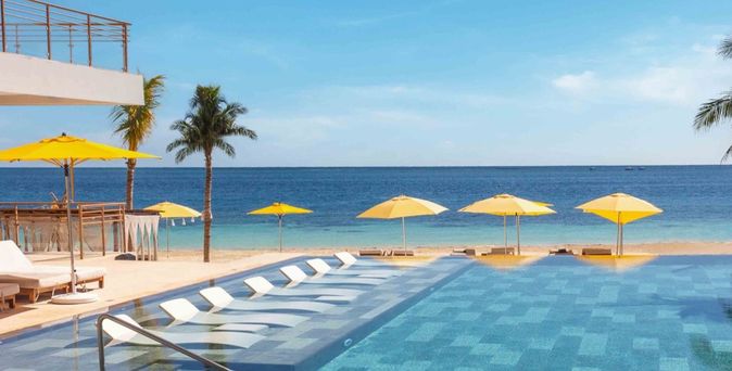 Pool mit Liegen und gelben Sonnenschirmen am Strand mit Palmen und Blick auf das Meer in Mexiko