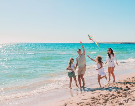 Familie mit zwei Kindern spielt am Strand und lässt einen bunten Drachen im Wind steigen, während das Meer im Hintergrund glitzert