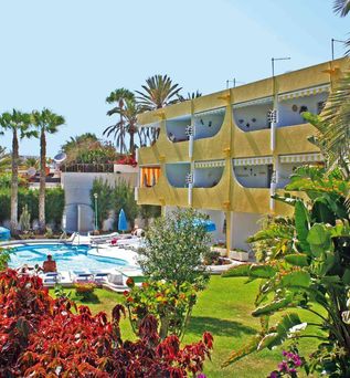 Gelbes Hotelgebäude mit Balkonen neben einem Pool, umgeben von grünen und roten Pflanzen, Palmen und blauem Himmel