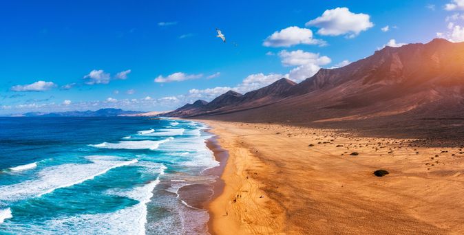 Langer Sandstrand an der Küste von Fuerteventura mit Wellen und Bergen im Hintergrund unter blauem Himmel