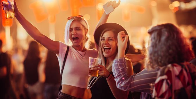 Drei junge Frauen feiern ausgelassen auf einer Party, halten Getränke in der Hand und lachen inmitten einer Menschenmenge mit bunter Beleuchtung