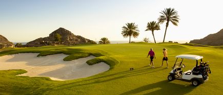 Zwei Personen stehen auf einem gepflegten Golfplatz mit Golfcart, Palmen und sandigen Bunkern, umgeben von Hügeln und Meerblick bei tiefstehender Sonne