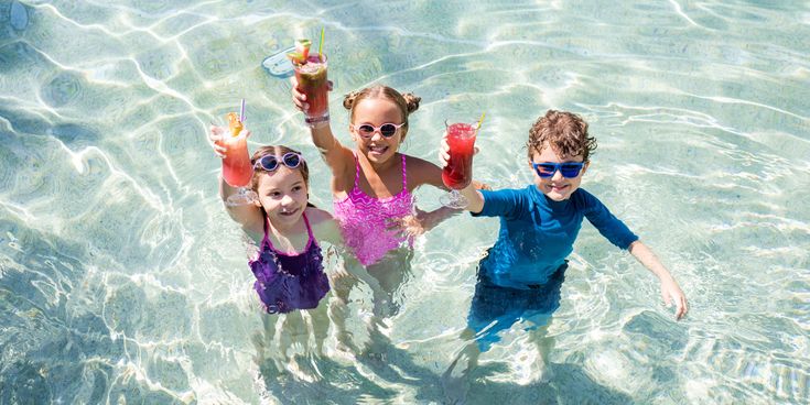 Drei Kinder stehen lachend im Pool und stoßen mit bunten Getränken in der Hand an