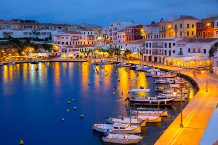 Abendstimmung am Hafen von Cales Fonts bei Mahón auf Menorca: beleuchtete Häuserfronten spiegeln sich im Wasser, kleine Fischerboote liegen am Kai