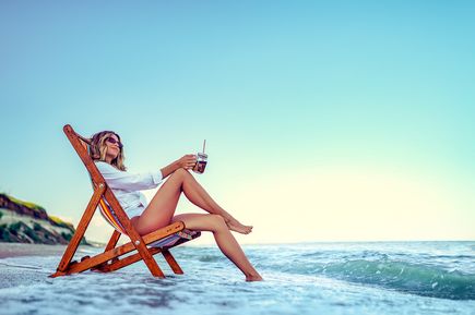 Frau sitzt entspannt in einem hölzernen Liegestuhl am Strand, hält ein Glas mit Strohhalm, Wellen im Hintergrund