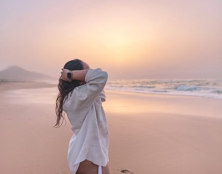 Frau mit langem Haar und weißem Hemd steht am Strand von Cofete auf Fuerteventura und blickt in den Sonnenuntergang.