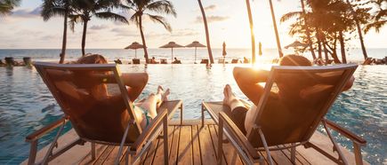 Zwei Personen liegen in Sonnenliegen am Pool und schauen auf das Meer sowie Palmen bei untergehender Sonne