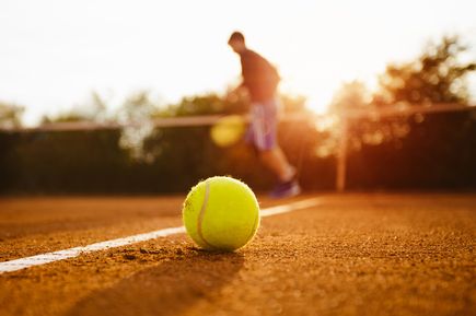 Nahaufnahme eines gelben Tennisballs auf der roten Sandplatz-Linie, unscharf im Hintergrund ein Spieler in Bewegung
