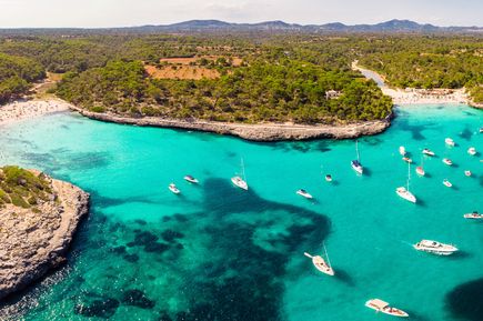 Luftaufnahme der Bucht Cala Mondragó auf Mallorca mit türkisblauem Wasser, Booten im Meer und von grünen Pinienwäldern umgebenen Stränden