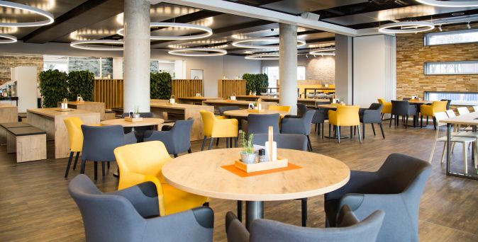 Moderne Firmenkantine mit runden Holztischen, gelben und grauen gepolsterten Stühlen, großen Säulen und runden Deckenleuchten