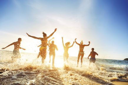 Gruppe junger Menschen rennt lachend und springend in die Meereswellen an einem sonnigen Strand, während das Wasser dynamisch aufspritzt.