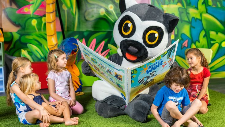 Katta-Maskottchen sitzt auf einem Teppich und liest mehreren Kindern aus einem Buch im bunten Indoor-Spielbereich mit Dschungelkulisse vor