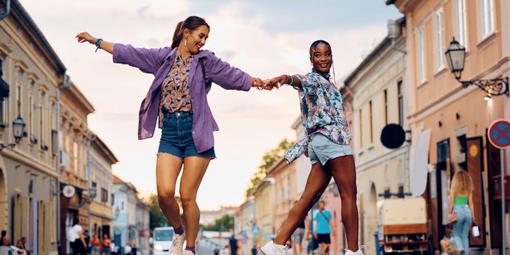 Zwei Frauen tanzen händehaltend durch eine Altstadtstraße mit historischen Gebäuden und urbanem Ambiente