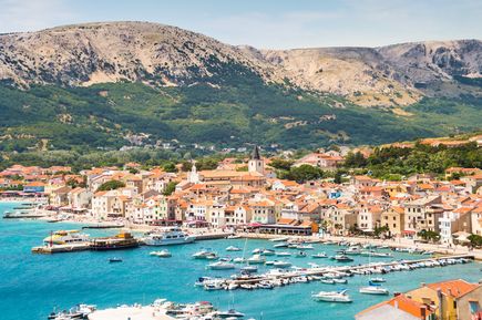 Panoramablick auf den Küstenort Baška mit Hafen, zahlreichen Booten, rot gedeckten Häusern und den kargen Bergen der Insel Krk im Hintergrund