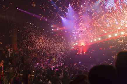 Große Menschenmenge feiert in einem Club mit bunter Beleuchtung, während von der Decke Konfetti herabfällt