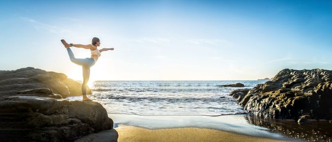 Frau übt eine Yoga-Pose auf Felsen am Strand, mit Blick auf das Meer und die aufgehende Sonne unter klarem Himmel