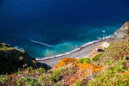 Blick von den Klippen von Ponta do Garajau auf Madeira auf einen schmalen Kiesstrand, das tiefblaue Meer und die steile Küstenlandschaft
