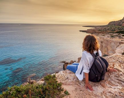 Frau mit schwarzem Rucksack sitzt auf Felsen und blickt auf das ruhige Meer bei Sonnenuntergang auf Zypern