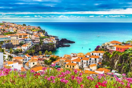 Blick über die Küstenstadt Câmara de Lobos auf Madeira mit orangefarbenen Dächern, steilen Felsen, türkisblauer Bucht, Segelbooten und blühenden rosa Oleandern im Vordergrund