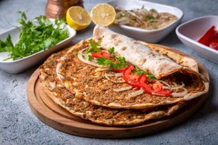 Lahmacun auf Holzbrett, belegt und gerollt mit Tomaten, Zwiebeln und Petersilie, daneben Zitronen und Kräuter
