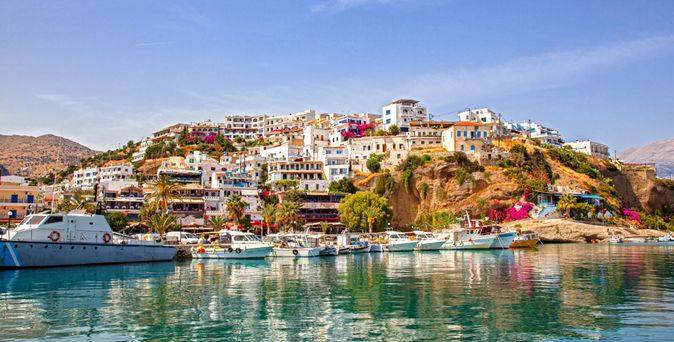 Hafen mit Booten vor einem Hügel mit weißen Häusern und blühenden Pflanzen auf Kreta